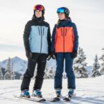 Mladý muž a žena v lyžiarskom oblečení stojaci na zasneženej hore so zjazdovkami a stromami.