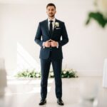 Muž v tmavomodrom obleku stojí v elegantnej svadobnej sieni s kvetinovou výzdobou v pozadí.