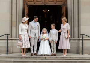 Rodina na prvej svätej omši pred kostolom, dievča v bielych šatách s venčekom, hostia v jarnom oblečení.