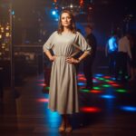 Žena v štýlových šatách stojí v modernom nočnom klube s farebnými svetlami na pozadí.