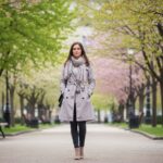 Žena v béžovom trenčkot a šále stojí v mestskom parku počas jarného dňa s rozkvitnutými stromami.