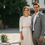 Svadobný pár pri civilnom obrade vonku, ženích v svetlom obleku, nevesta v bielych šatách so svadobnou kyticou.