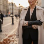 Žena v béžovom trenčkot s čiernymi koženými rukavicami pózuje na mestskej ulici počas zlatého svetla